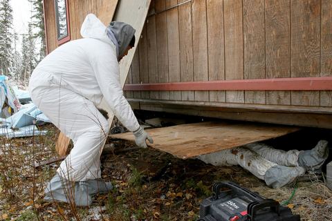 Worker in white coveralls lifting a plywood skirt panel to reach insulation under a raised house