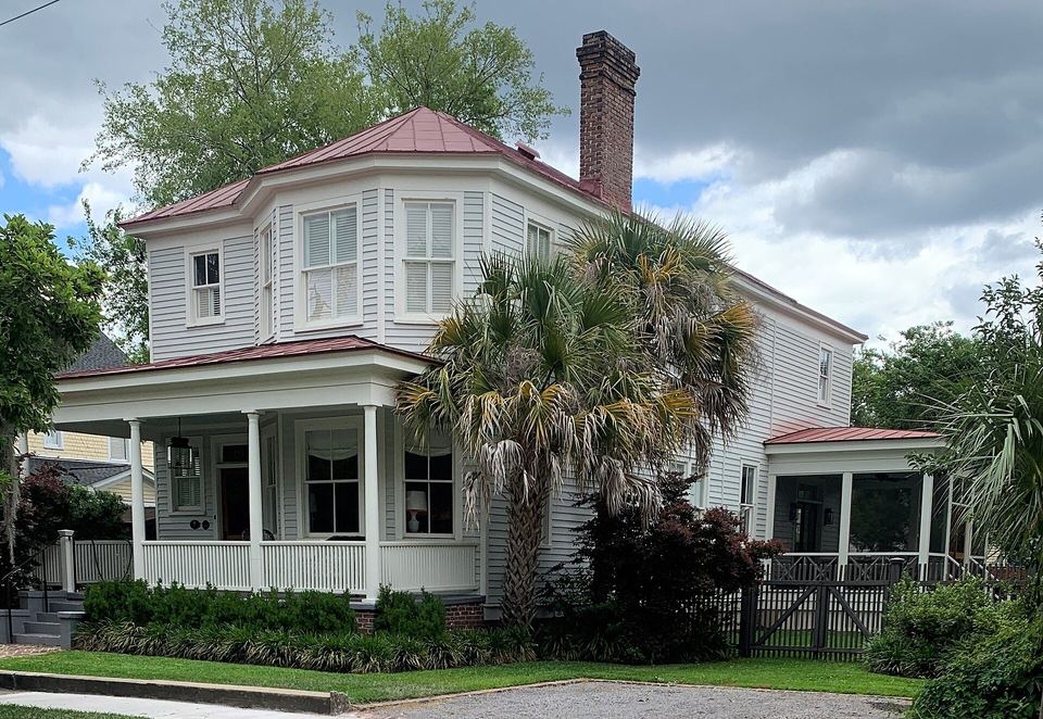 Two-story white clapboard house with a wraparound porch, metal roof and palmetto trees